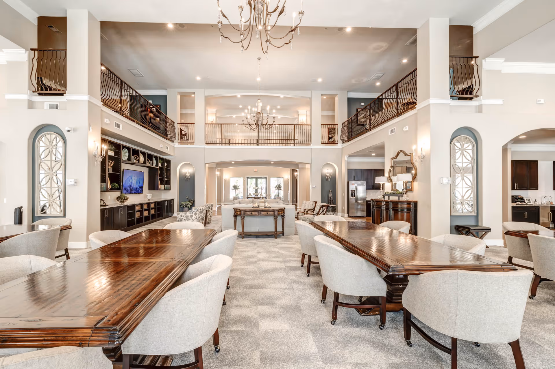 Spacious and elegant common area in a senior living facility featuring two long wooden tables with beige upholstered chairs, a sitting area with sofas and armchairs, a large flat-screen TV mounted on a dark built-in shelving unit, and a mezzanine balcony with decorative railings overlooking the room. The room is well-lit with chandeliers and wall sconces, and has a neutral color palette with light walls and carpeted floors.