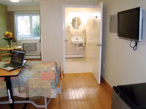Senior living bedroom with bed, table, wall-mounted TV, and doorway to a bathroom.