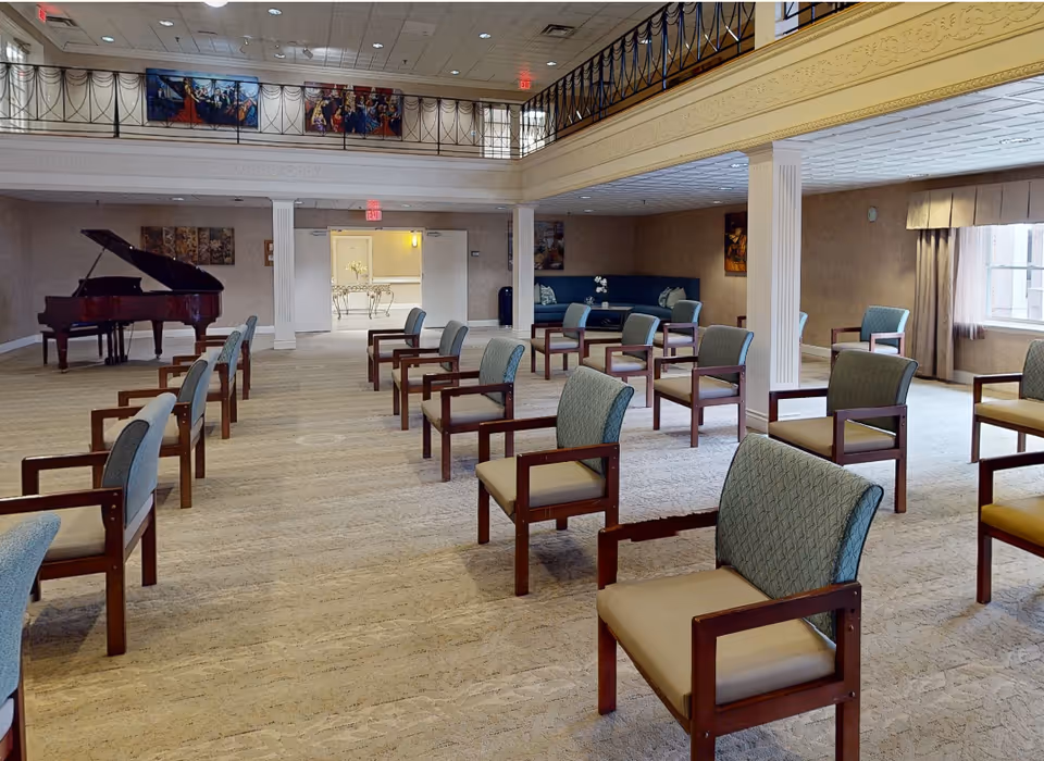 Large airy common room in a senior living facility with spaced armchairs, a grand piano, and an upper gallery.