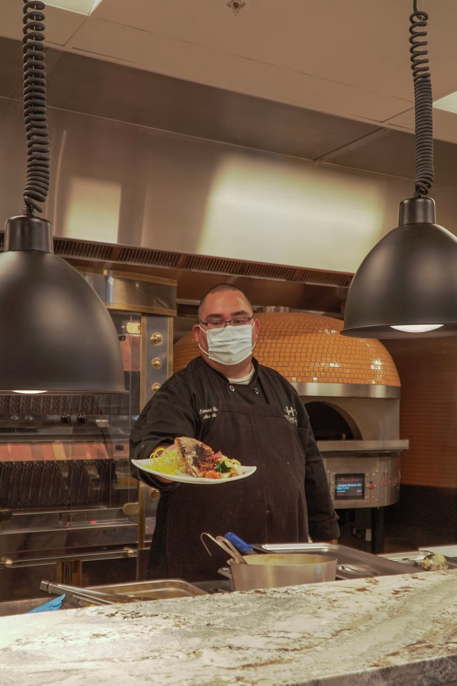 A chef wearing a black uniform and a face mask is standing in a kitchen holding a plate of food. The kitchen has a large oven with a tiled dome and two black hanging lamps above the counter.