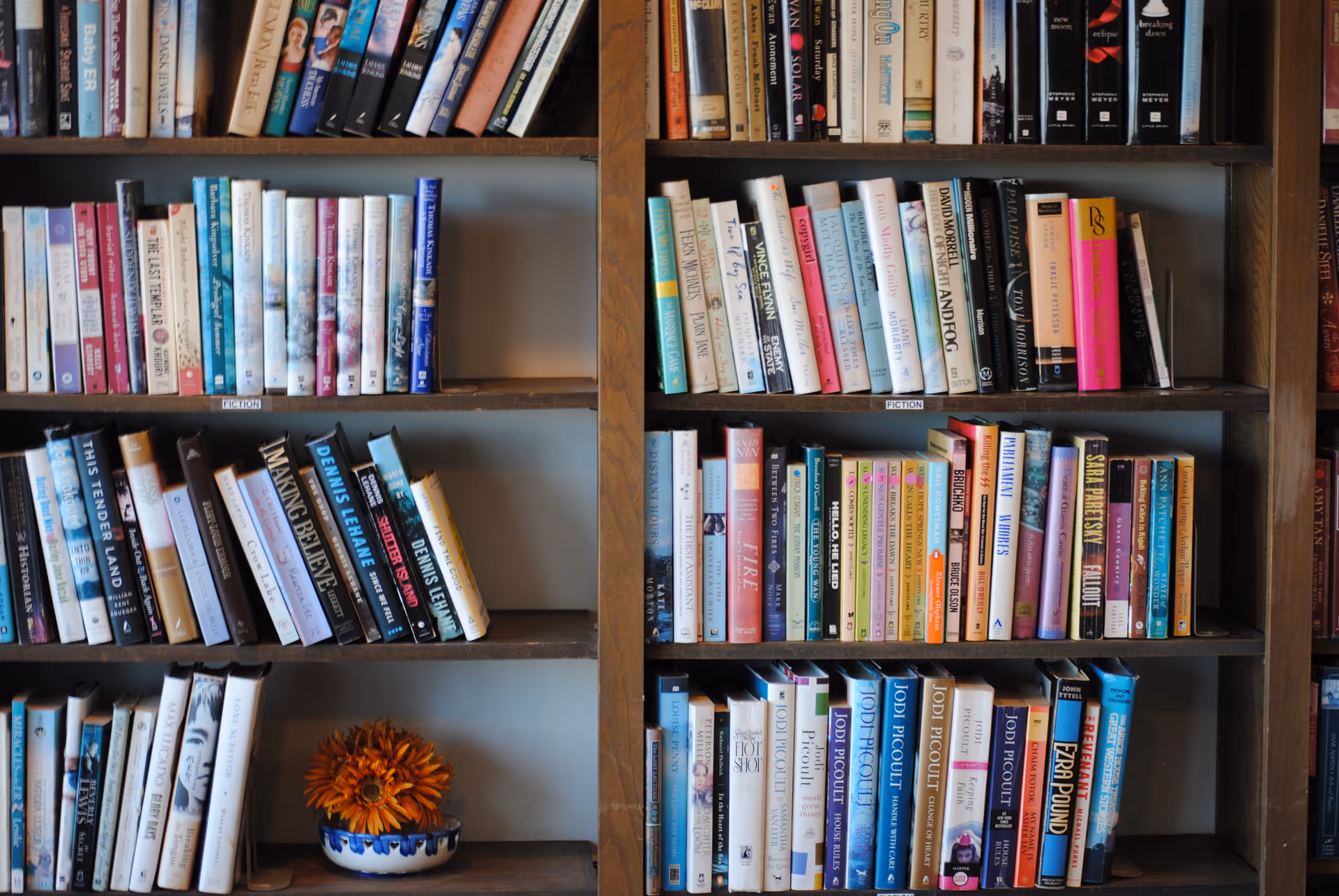 Wooden bookshelf filled with assorted books and a small bowl of orange flowers on a lower shelf.