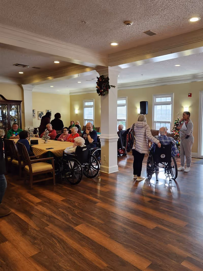 A group of elderly people, some in wheelchairs, gathered in a spacious room with wooden floors and cream-colored walls. Several people are seated around a large table covered with a yellow tablecloth, while others are standing or sitting near windows. The room is decorated with a small Christmas tree and a wreath on a pillar, indicating a festive atmosphere.