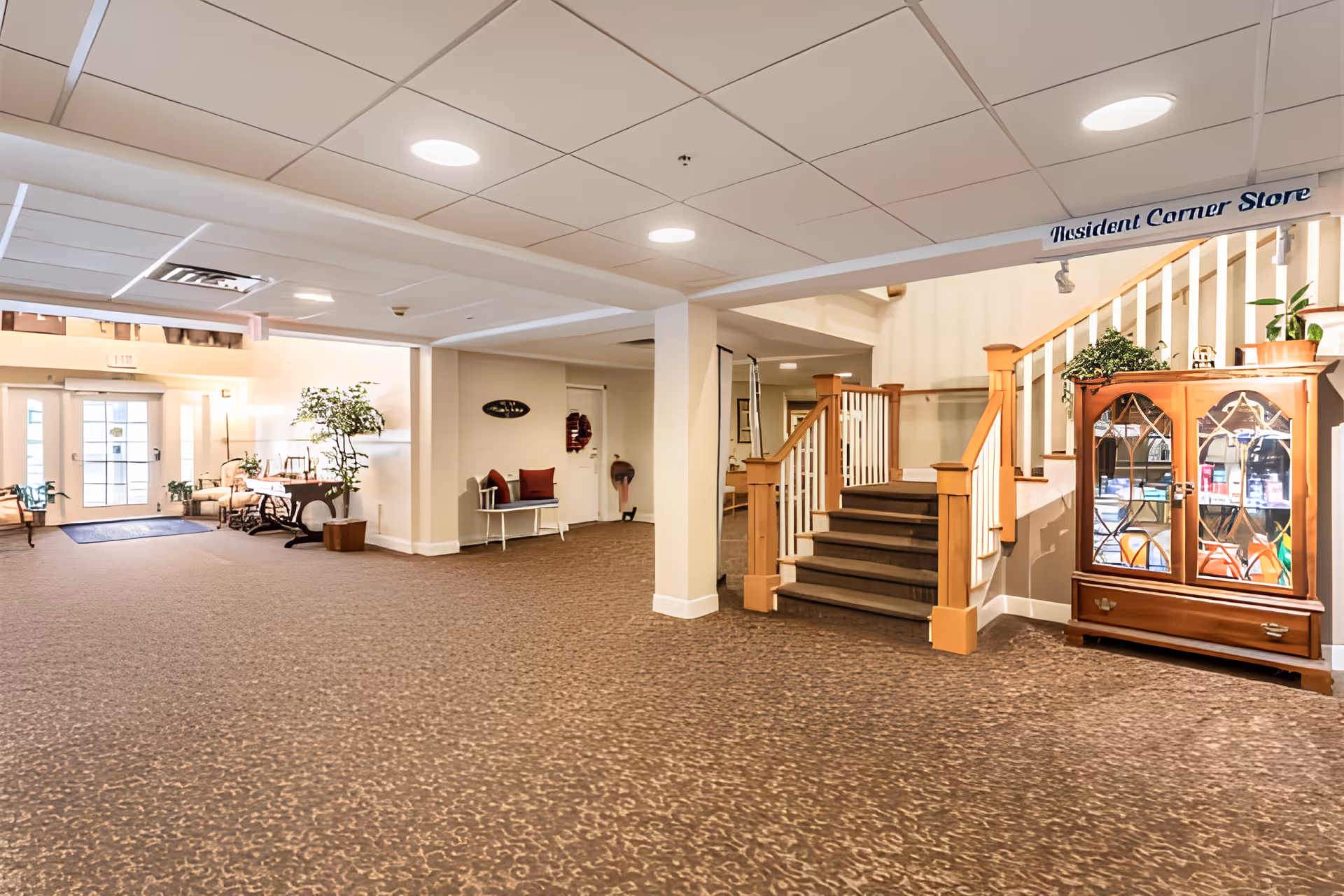 Spacious interior common area with carpeted floor, a staircase with wooden railings, a glass-front cabinet with plants and items inside, a bench with red cushions, and a table with plants near the entrance door.