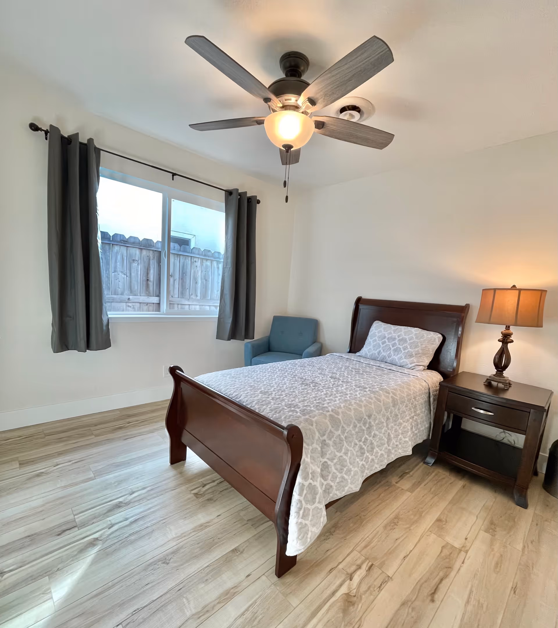 A bedroom with a single wooden bed covered with a light gray patterned bedspread and a matching pillow. Next to the bed is a dark wooden nightstand with a lamp on it. A blue armchair is placed near a window with dark gray curtains. The room has light wood flooring and a ceiling fan with a light fixture.