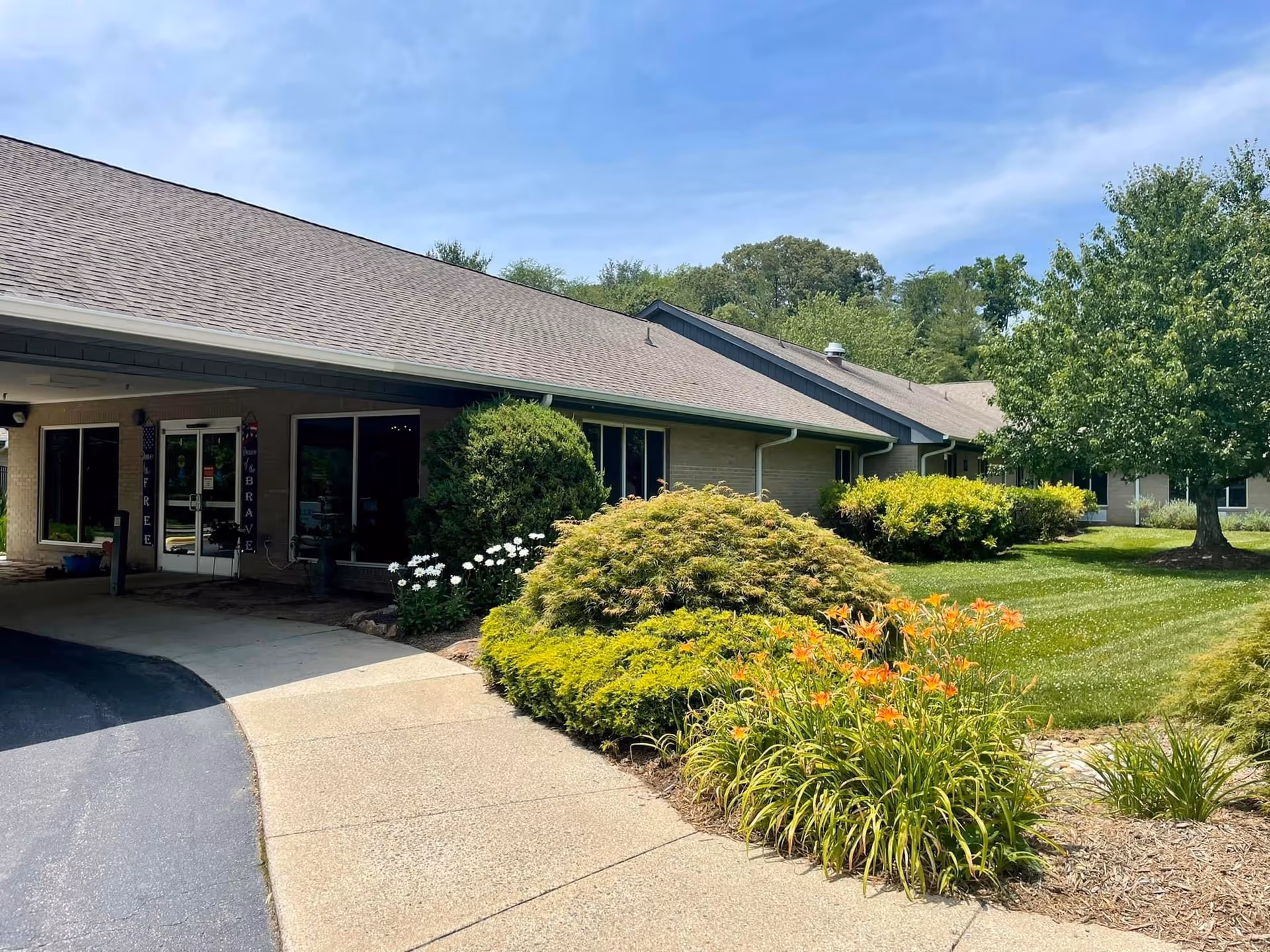 Front entrance of a single-story building with a covered drop-off, walkway, and landscaped shrubs and flowering plants.