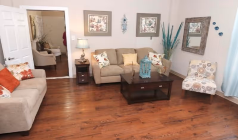 A cozy living room with wooden flooring, featuring two beige sofas with colorful cushions, a patterned armchair, a dark wooden coffee table with decorative items, a side table with a lamp, framed artwork on the walls, a large mirror, and a vase with tall blue-green decorative branches.