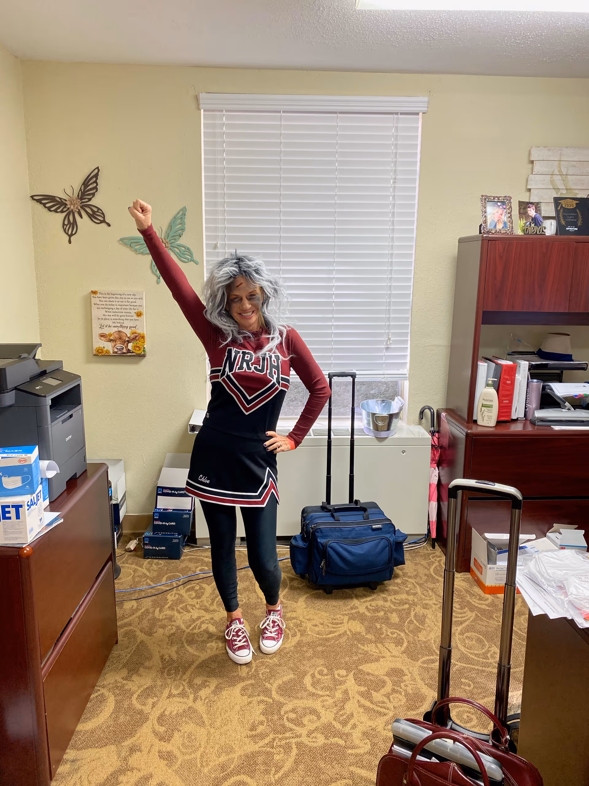A woman with gray hair wearing a maroon and black cheerleader outfit stands in an office room with one arm raised and smiling. The room has beige walls, a window with closed blinds, a wooden cabinet with framed photos and books, a printer, boxes, and two rolling suitcases on a patterned carpet.