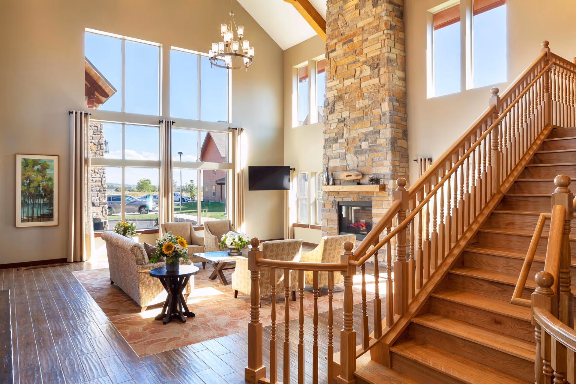 A bright and spacious living room area with large floor-to-ceiling windows letting in natural light. The room features a stone fireplace, a wall-mounted TV, comfortable seating including sofas and armchairs, a wooden staircase with railings leading upstairs, and a small round table with a vase of sunflowers. The floor is wooden with a patterned area rug, and there is a framed painting on the wall.