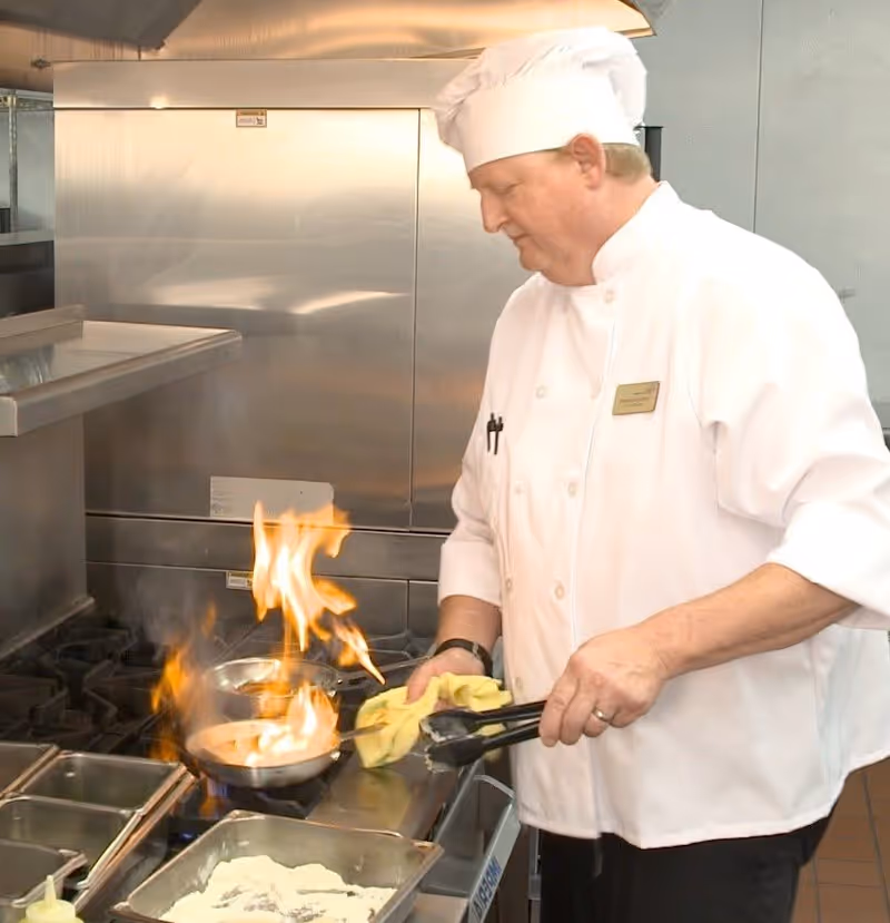 A chef wearing a white uniform and hat is cooking in a commercial kitchen, holding tongs and a yellow cloth while flames rise from a pan on the stove.