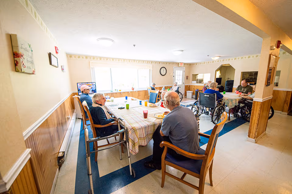 A group of elderly people, some in wheelchairs, sitting around two tables with checkered tablecloths in a bright dining room. They are eating and drinking, with cups and plates on the tables. The room has light-colored walls with wood paneling, a large window, and a clock on the wall.