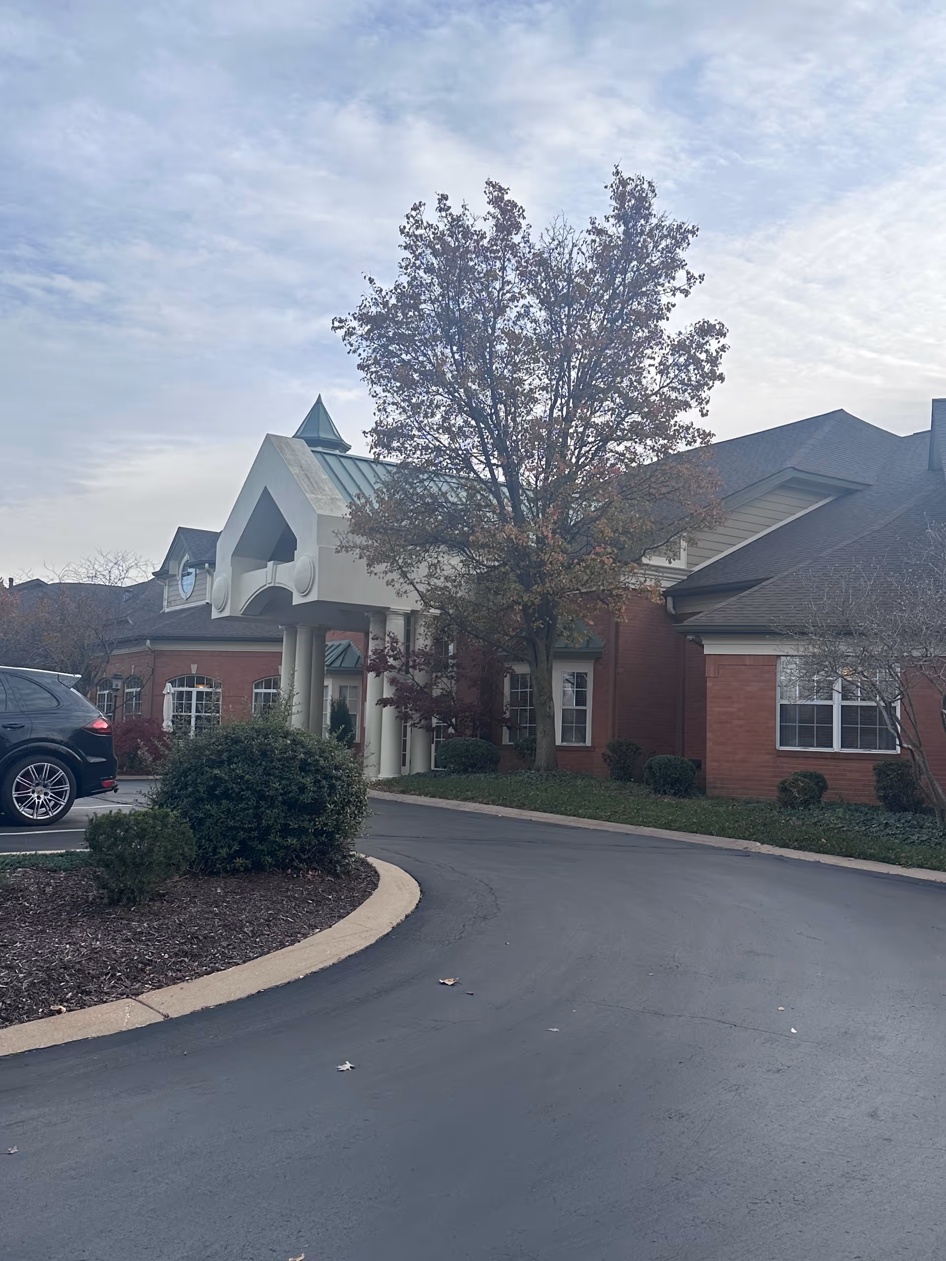 Exterior view of McKnight Place Assisted Living & Memory Care building with a curved driveway, a tree with autumn leaves, and some bushes in front. A black car is parked on the left side near the building entrance.