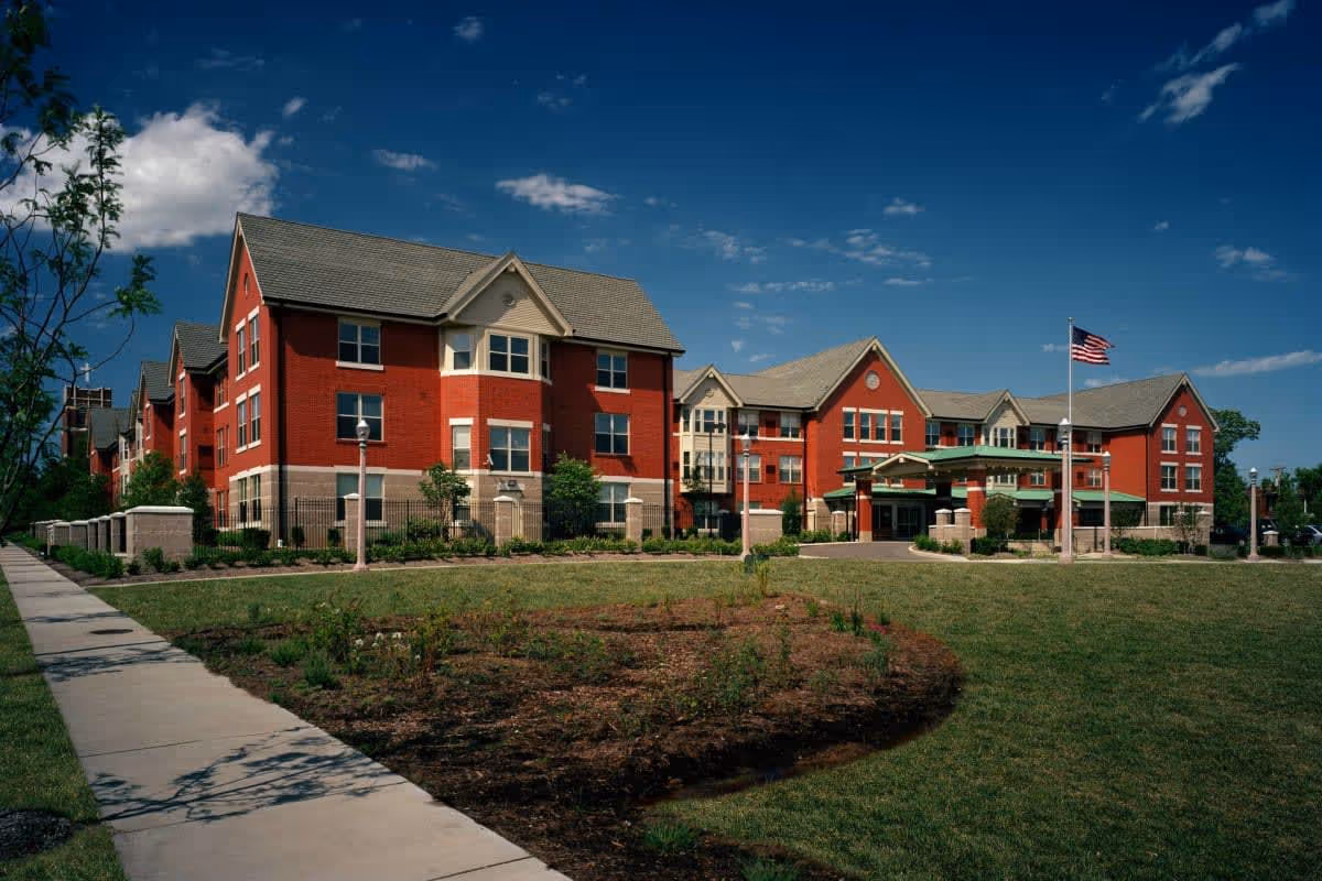 Exterior view of McCormack House-Forest Park Southeast, a large red brick building with multiple windows and a gray roof, surrounded by a green lawn and a sidewalk. An American flag is flying on a flagpole near the entrance under a covered driveway.