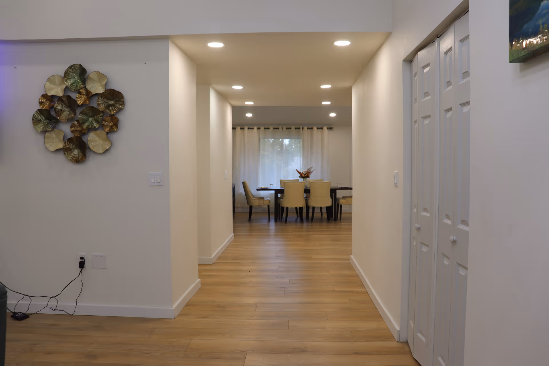 View down a hallway with wooden flooring leading to a dining area with a table and six chairs. The walls are painted white, with recessed ceiling lights illuminating the space. On the left wall, there is a decorative metal wall art piece, and on the right side, there are white closet doors. A window with curtains is visible in the dining area.