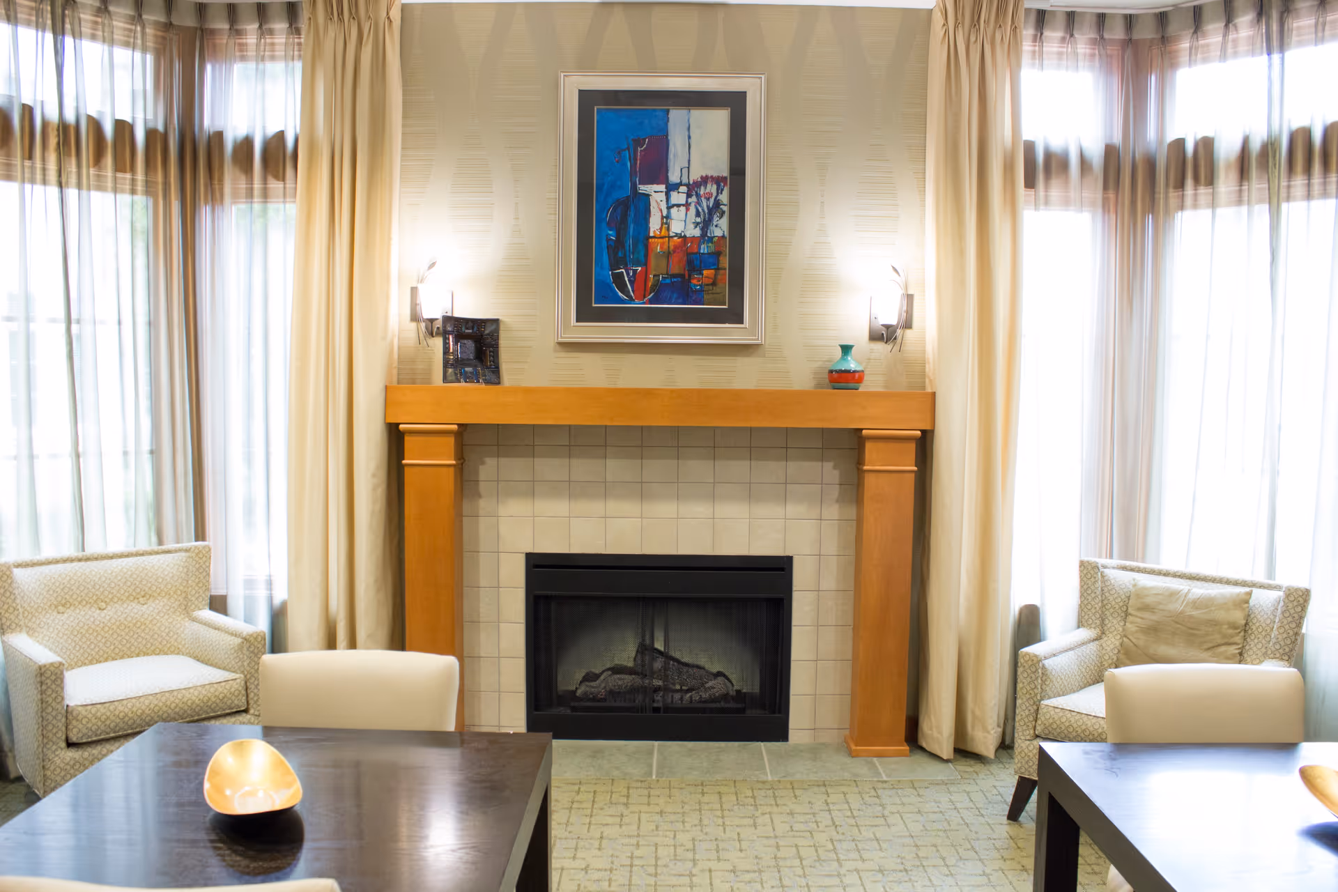 A cozy living room area with a fireplace centered on a tiled wall and wooden mantel. Above the mantel hangs a colorful abstract painting. On either side of the fireplace are wall-mounted lights and small decorative items on the mantel. Two armchairs with patterned upholstery are positioned near large windows with sheer curtains, and a dark wooden table with a decorative bowl is in the foreground.