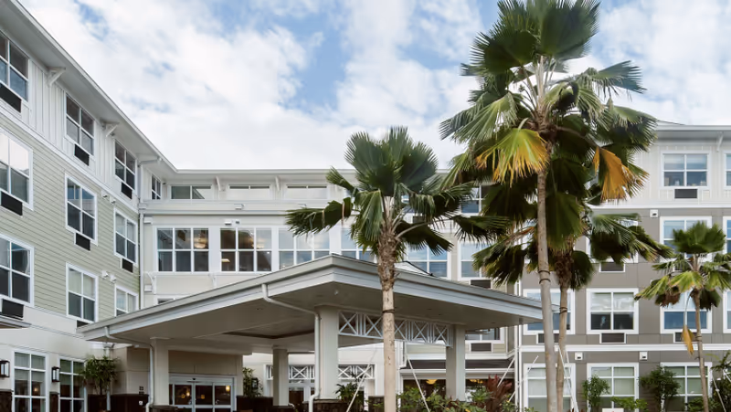 Front entrance of a multi-story senior living building with a covered porte-cochère and palm trees.