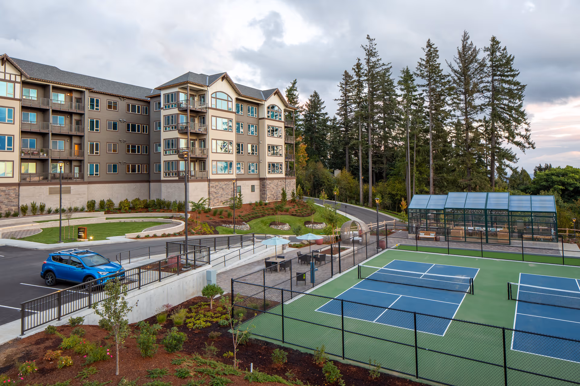 View of a senior living facility named Touchmark in the West Hills featuring a multi-story residential building, a parking area with a blue car, landscaped gardens, and outdoor pickleball courts enclosed by a fence. Tall trees and a partly cloudy sky are visible in the background.