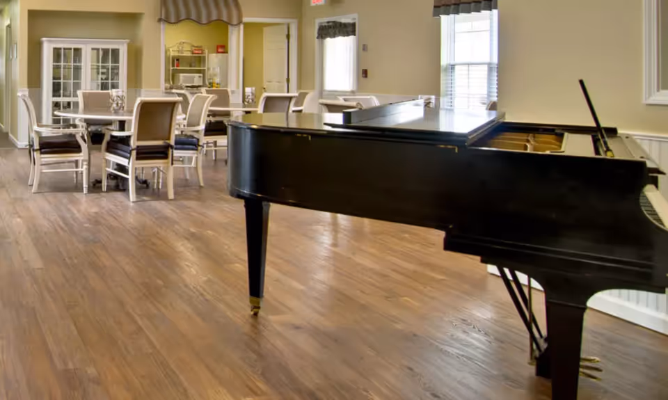 A spacious senior living common room with a black grand piano in the foreground and round dining tables and chairs in the background.