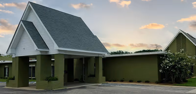 Exterior view of a single-story building with a green facade and a white peaked roof entrance, set against a partly cloudy sky during sunset.