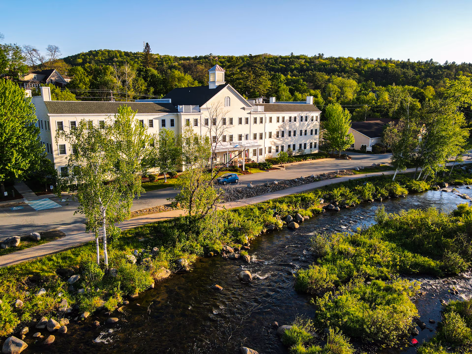 A large white building along a river, surrounded by trees and greenery.