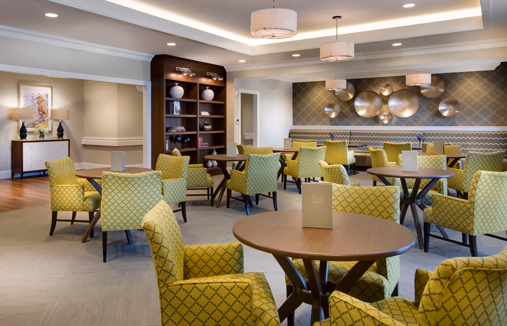 A well-lit dining room with multiple round wooden tables surrounded by yellow patterned upholstered chairs. The room features modern pendant lights, a decorative wall with circular metallic art pieces, a built-in wooden bookshelf with decorative items, and a sideboard with lamps and artwork. The flooring is a combination of carpet and wood.