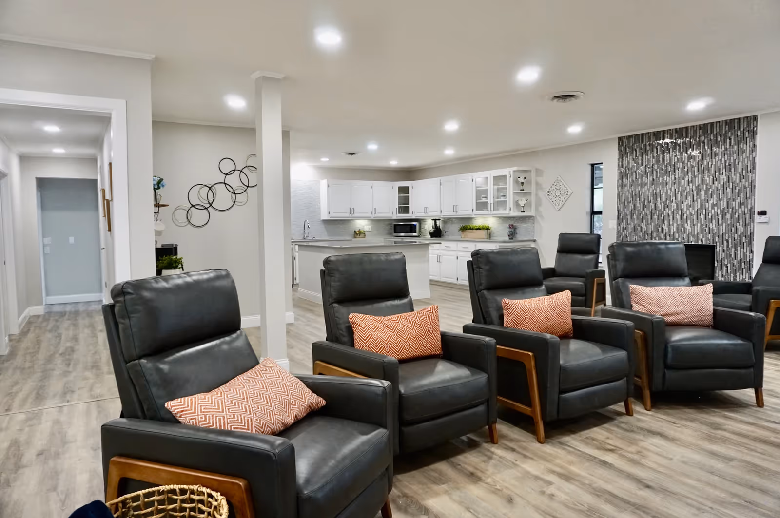 Bright communal living area with a row of dark leather recliners with orange patterned pillows facing an open white kitchen.