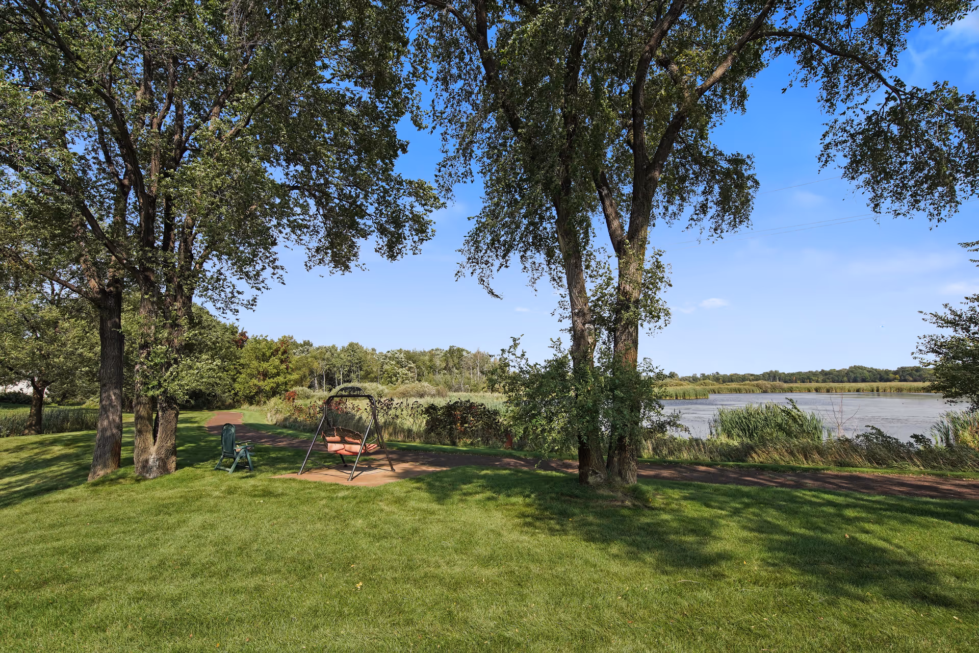 A peaceful outdoor scene at Keller Lake Commons featuring green grass, several tall trees, a swing bench, and a single chair near a walking path. In the background, there is a lake with reeds and a clear blue sky.