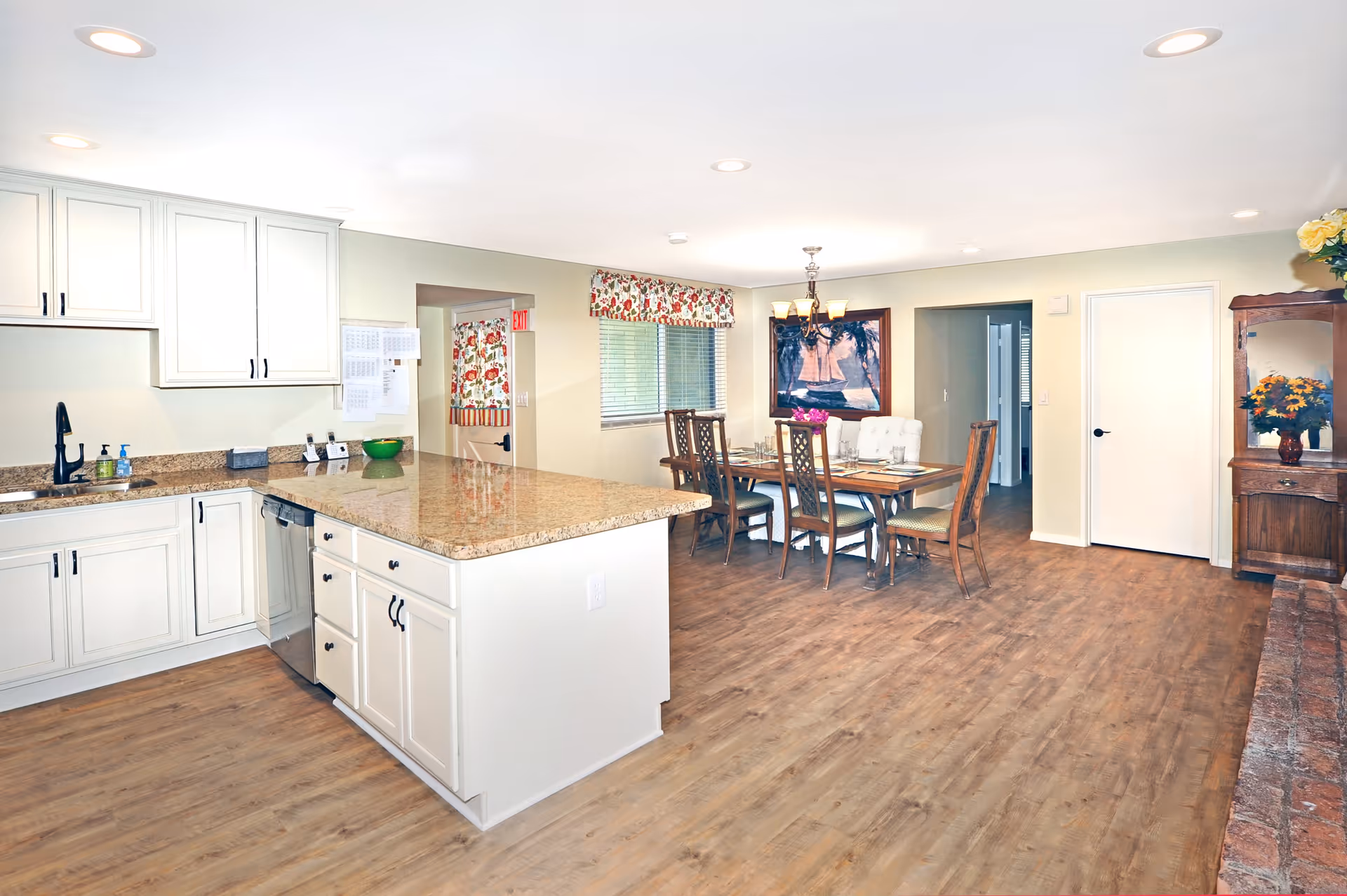 Bright and spacious kitchen and dining area in an assisted living facility. The kitchen features white cabinets, granite countertops, a black faucet, and a dishwasher. The dining area has a wooden table set with six chairs, a chandelier overhead, a window with floral curtains, and a framed painting on the wall. There is a wooden cabinet with a mirror and a vase of flowers on the right side of the room. The floor is wood-style laminate.