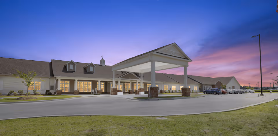Exterior view of a single-story senior living facility building at dusk with a covered entrance, several windows lit from inside, a parking lot with cars, and a purple and pink sky in the background.