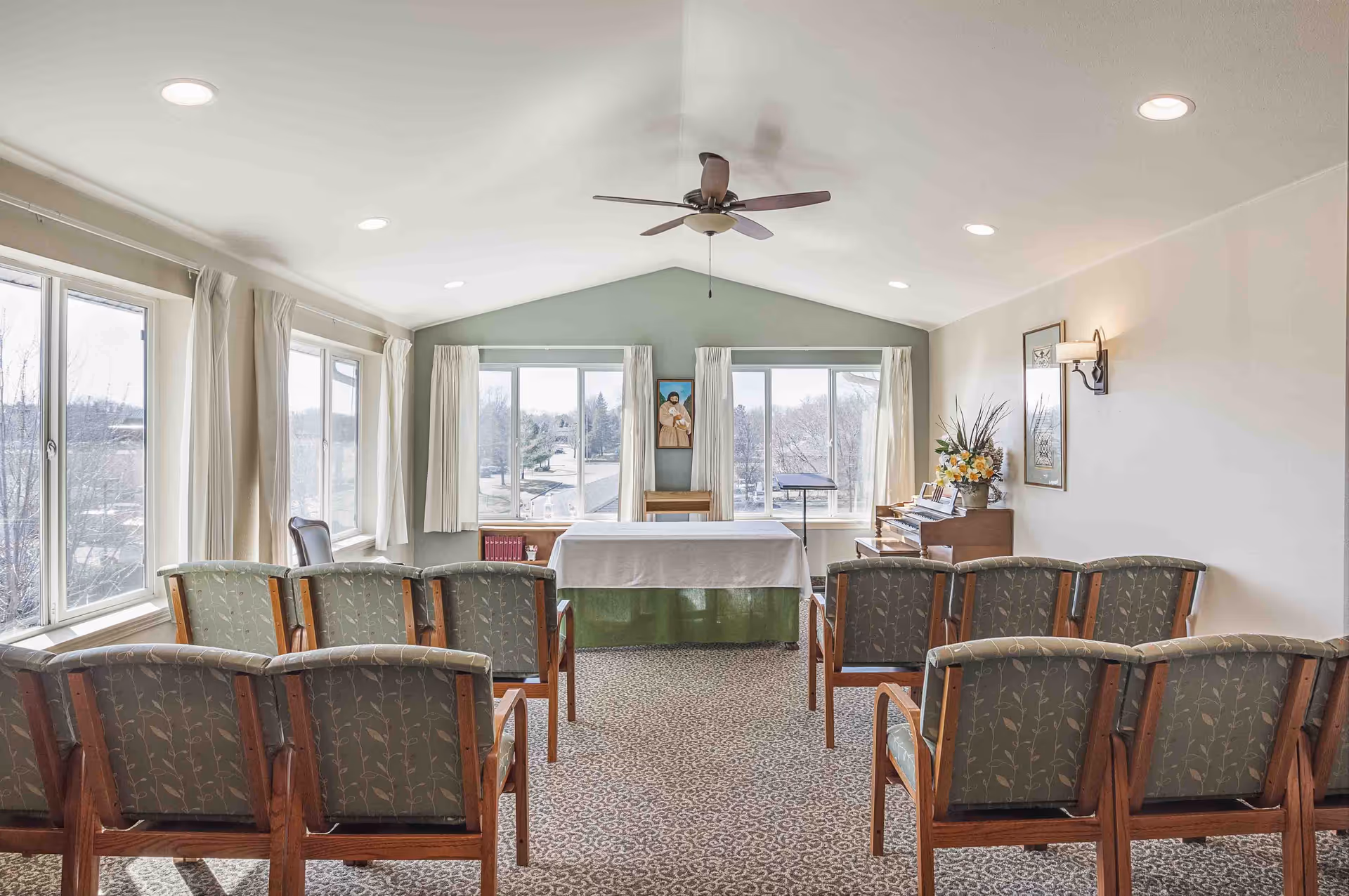 Chapel-style common room with rows of upholstered chairs facing a covered table/altar, large windows, and a piano.