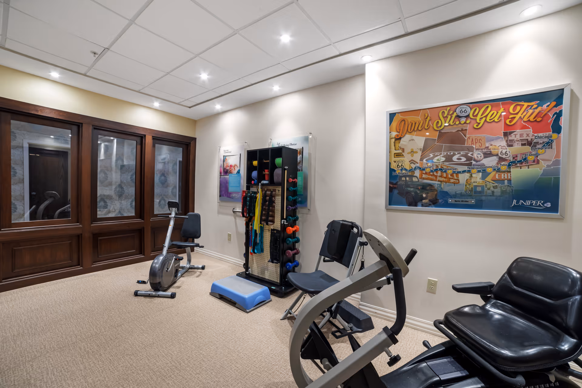 A small fitness room with exercise equipment including a recumbent bike, a seated leg press machine, and a rack holding various dumbbells, resistance bands, and exercise mats. The room has beige carpet, cream-colored walls, recessed ceiling lights, and a large framed poster on the wall that says 'Don't Sit... Get Fit!' with a Route 66 theme. There are large wooden-framed windows on one side.