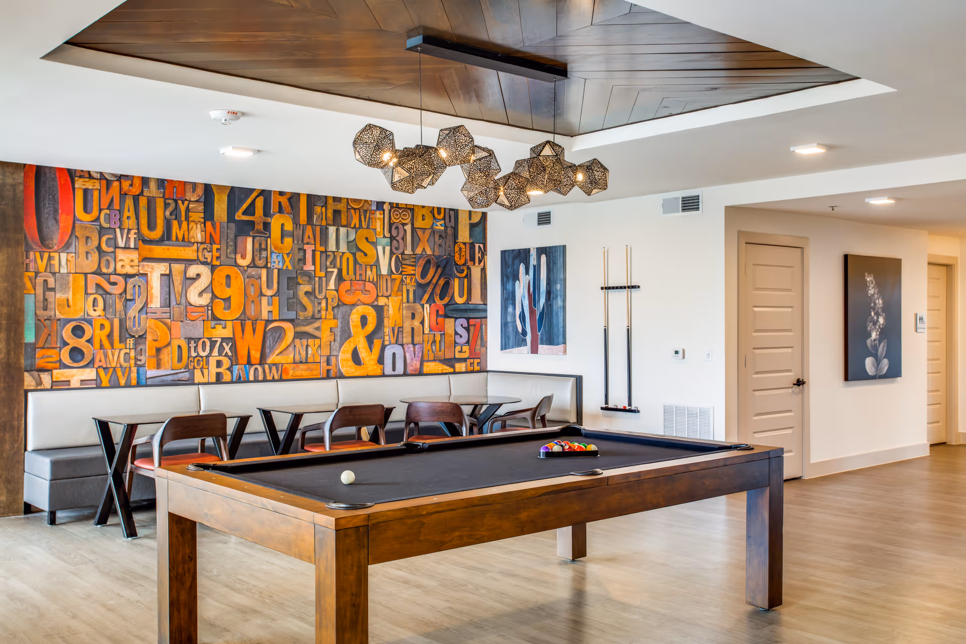 A modern recreational room featuring a wooden pool table with a black felt surface, a set of pool balls arranged in a triangle, and a cue ball. Behind the pool table is a seating area with a long cushioned bench and several wooden chairs around tables. The wall behind the seating area is decorated with a colorful abstract mural of letters and numbers. The ceiling has a wooden inset with a geometric pendant light fixture. The room has light-colored walls and flooring, with two closed doors and framed artwork on the walls.