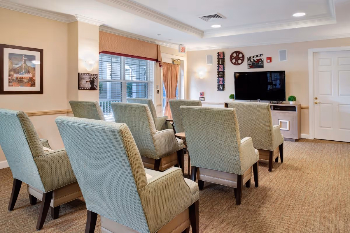 A bright community sitting room with upholstered chairs arranged in rows facing a flat-screen TV and wall decor.
