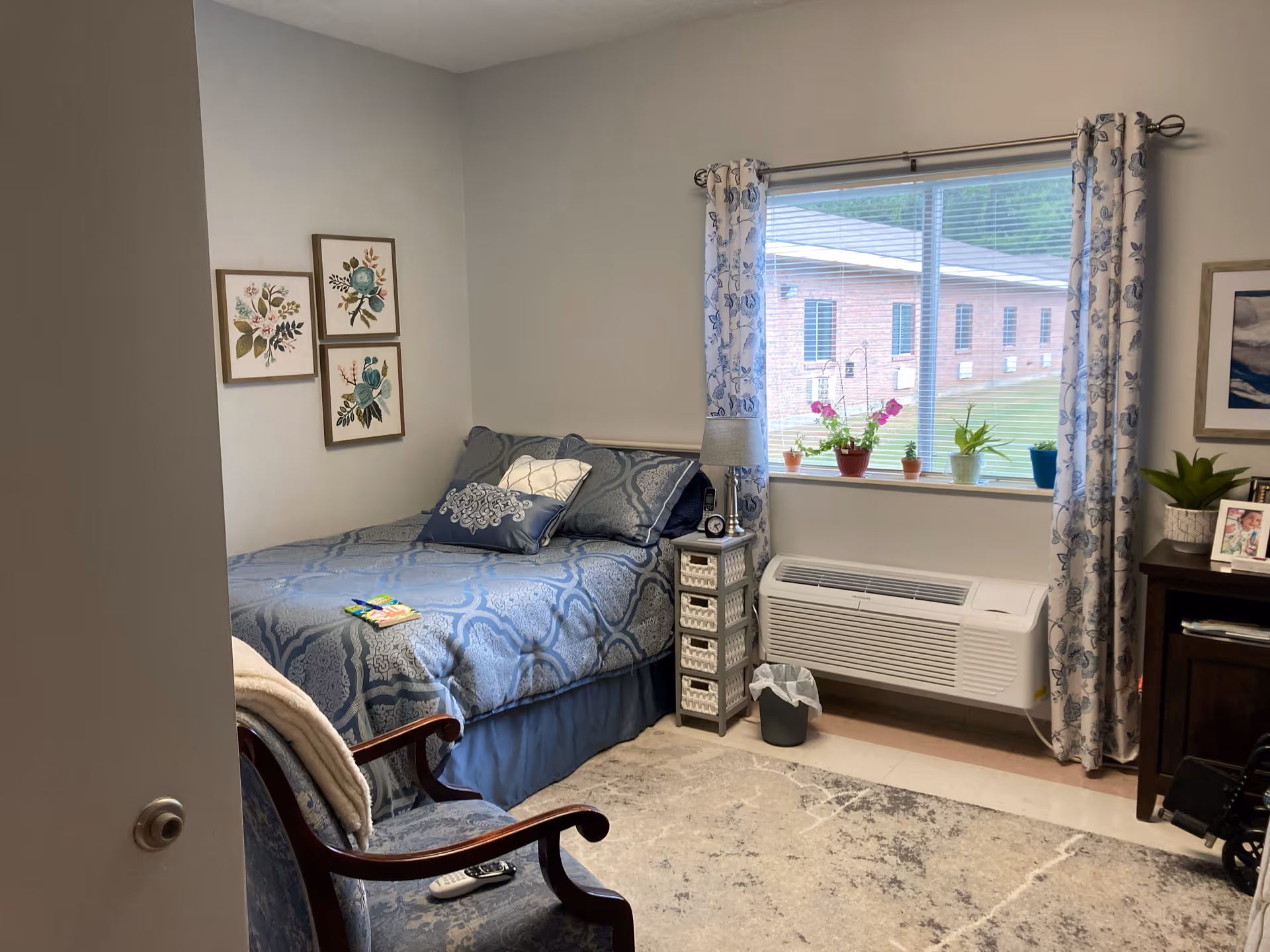 A cozy bedroom with a blue patterned bedspread and matching pillows on a bed against the wall. There are three framed floral artworks hanging above the bed. A small bedside table with a lamp, clock, and small drawers is next to the bed. A window with floral curtains shows a view of a brick building outside. Several potted plants are placed on the windowsill. A cushioned armchair with a remote control on it is in the foreground, and a dark wooden dresser with framed photos and a plant is on the right side of the room. The floor has a light-colored rug with a subtle pattern.