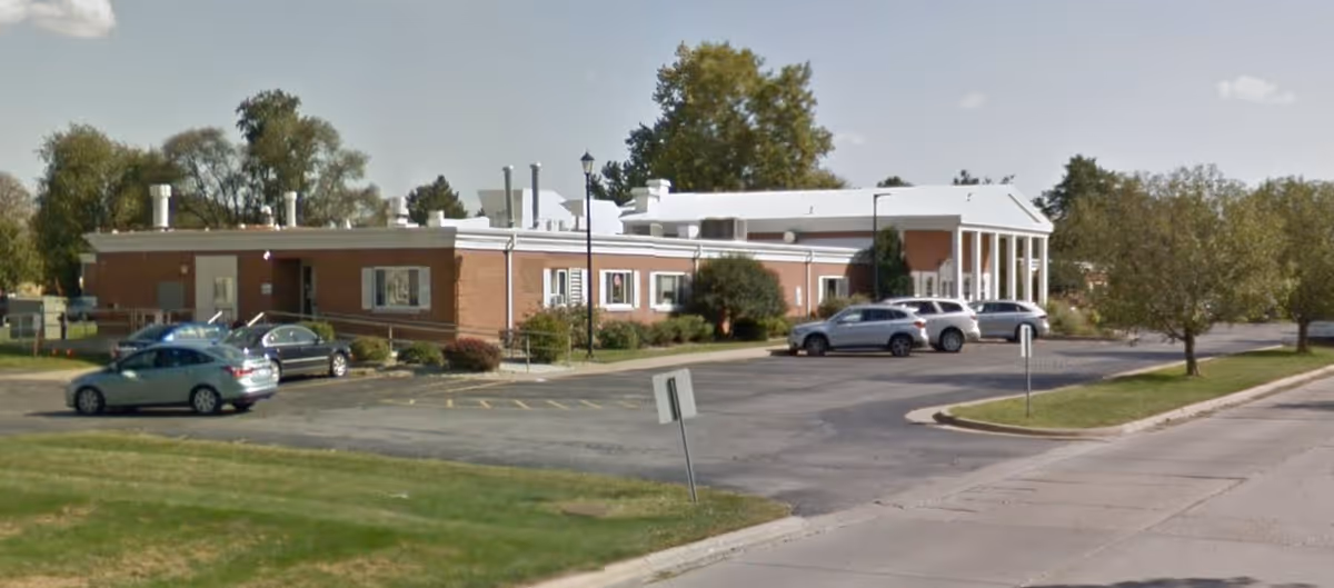 Brick single-story senior care facility with a columned entrance, parked cars, and a paved parking lot under a partly cloudy sky.