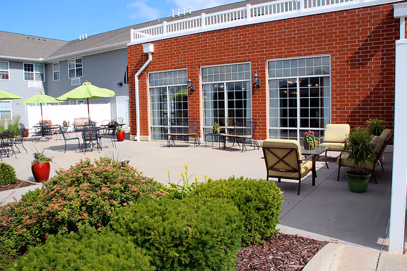 Outdoor patio area at John M. Evans Supportive Living Community featuring several metal tables with green umbrellas, cushioned chairs around a glass coffee table, potted plants, and a red brick building with large windows and white trim in the background.