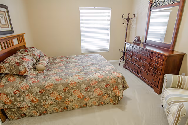 A bedroom with a floral patterned bedspread on a wooden bed frame, a wooden dresser with a large mirror, a coat rack, and a striped armchair. A window with closed blinds is on the wall behind the bed.