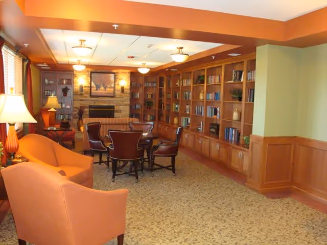 A cozy living room area with orange armchairs, a round table surrounded by four dark wooden chairs, and a large built-in wooden bookshelf filled with books and decorative items. The room features a fireplace with a framed picture above it, warm lighting from ceiling fixtures and table lamps, and carpeted flooring with a patterned design.