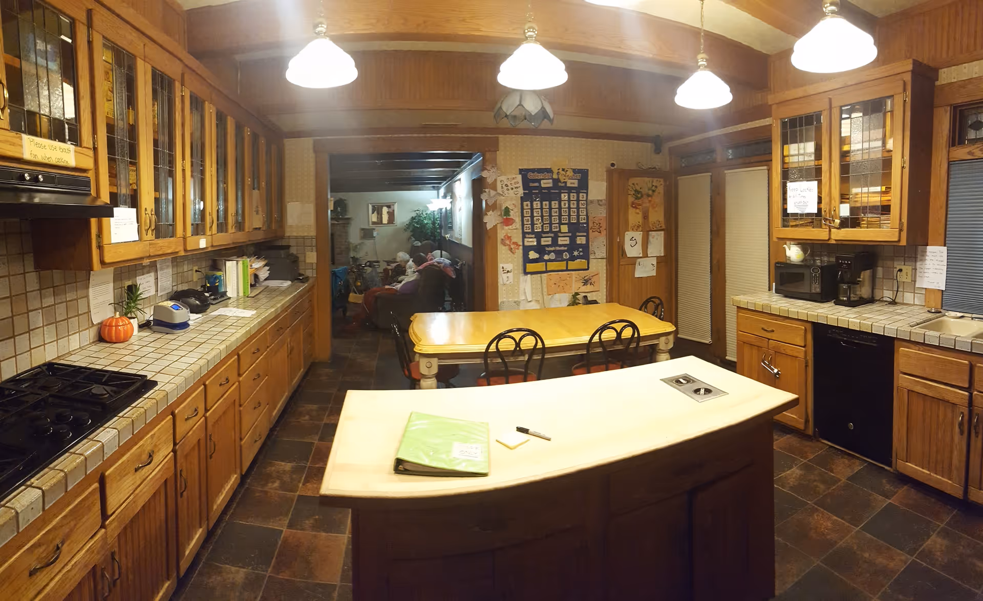 Communal kitchen with wood cabinetry, tiled countertops, a central island and a dining table in the background.