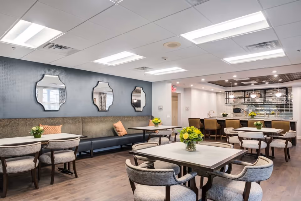 A bright and modern dining area with several tables and cushioned chairs arranged neatly. The room features a long bench along one wall with decorative pillows and three uniquely shaped mirrors above it. In the background, there is a kitchen area with a countertop and bar stools, pendant lights hanging from the ceiling, and a tiled backsplash. Fresh flower arrangements are placed on each table, adding a touch of color to the space.