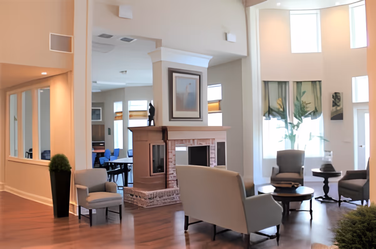 A bright and spacious living room area in a senior living facility with wooden flooring, a central fireplace with a framed picture above it, several armchairs and small tables arranged around the room, large windows with sheer curtains allowing natural light to fill the space, and a potted plant near the entrance.