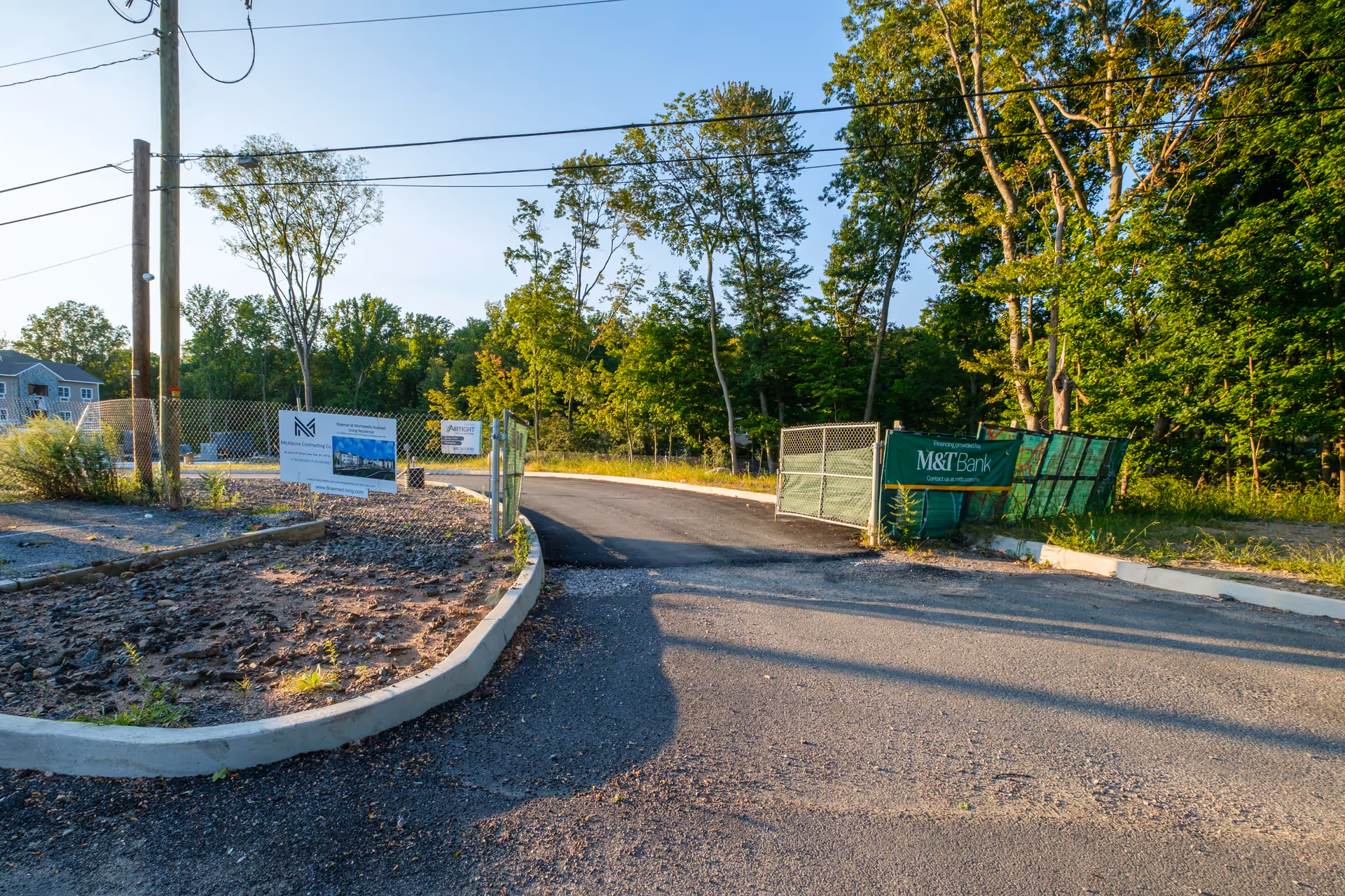 Entrance to a construction site with a paved road leading into a fenced area surrounded by trees and greenery. Several signs are posted on the fence, including one for M&T Bank and another for McAlpine Contracting Co. The sky is clear and the scene is lit by sunlight.