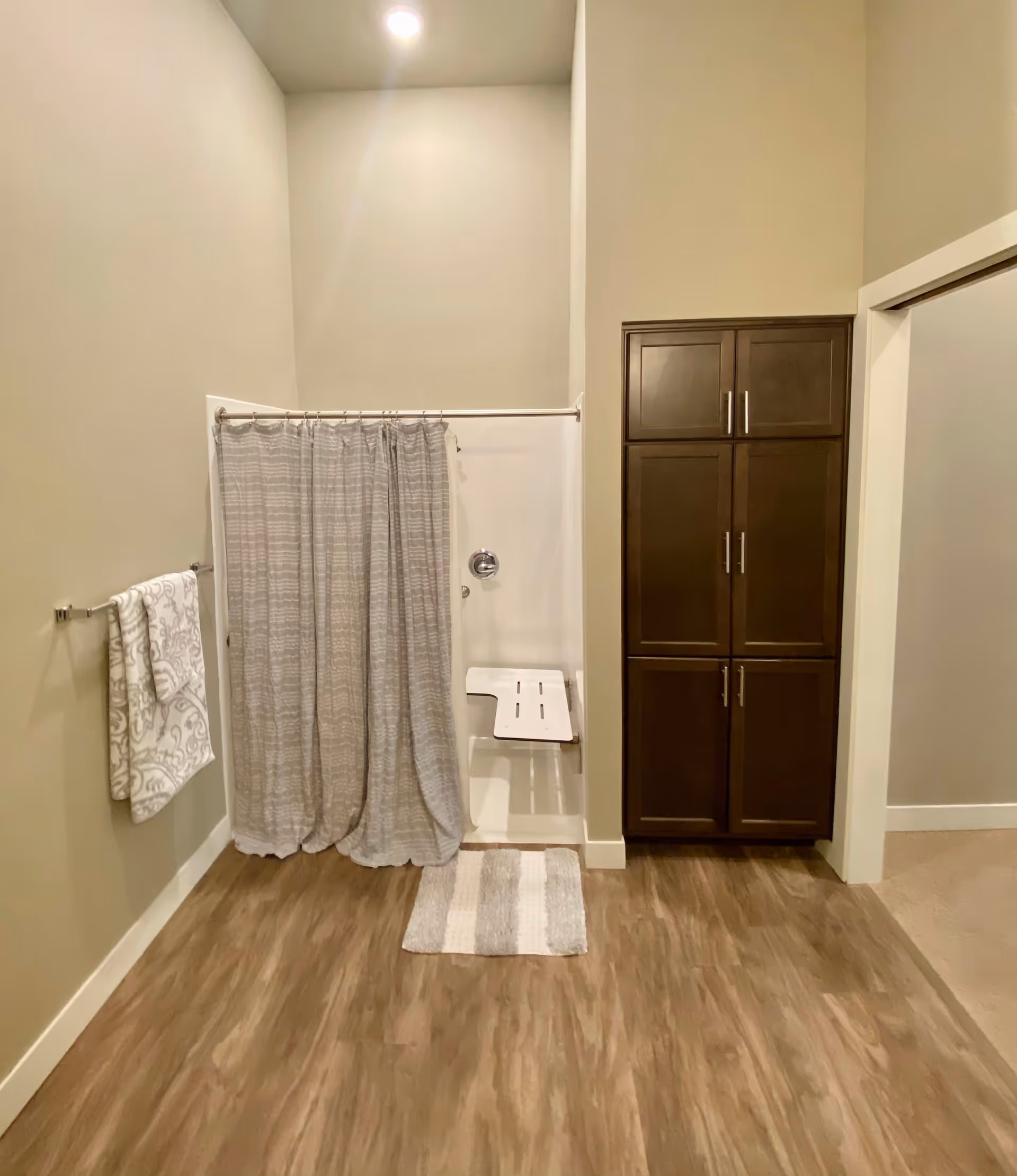 A bathroom with a walk-in shower that has a gray patterned shower curtain and a white fold-down shower seat. There is a towel rack on the left wall with two patterned towels hanging on it. To the right of the shower is a tall dark brown cabinet with multiple doors. The floor has wood-like vinyl flooring and a small striped bath mat in front of the shower.