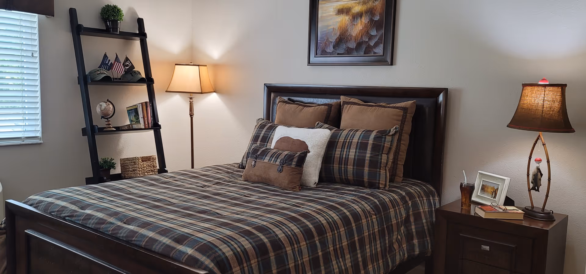 A cozy bedroom with a dark wooden bed frame and a plaid bedspread in shades of brown and gray. The bed is adorned with multiple pillows in coordinating colors. To the right of the bed is a wooden nightstand with a lamp, a framed photo, a glass with a straw, and a book. On the left side of the room, there is a black ladder-style shelf holding decorative items including small plants, hats, flags, a globe, and books. A floor lamp stands next to the shelf, and a window with blinds is partially visible on the left wall. A framed artwork hangs above the bed on a light-colored wall.