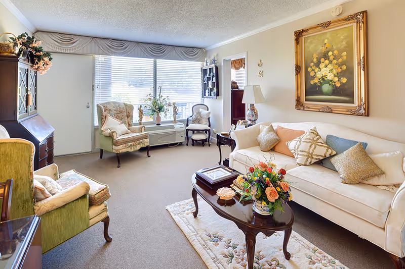 A cozy living room with a cream-colored sofa adorned with multiple decorative pillows, a wooden coffee table with a floral arrangement, two upholstered armchairs, a wooden cabinet, and a large window with blinds letting in natural light. A framed painting of yellow flowers hangs on the wall above the sofa.