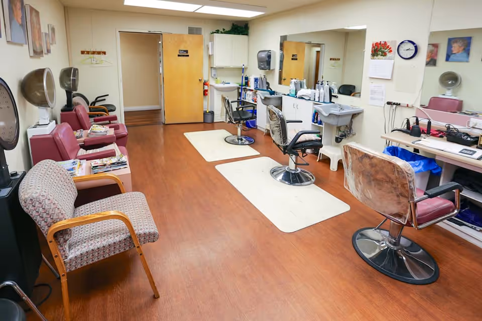 Interior view of a senior living facility's hair salon with wooden flooring, several salon chairs, hair drying stations, mirrors, and a waiting area with cushioned chairs. The room is well-lit with fluorescent ceiling lights and has various hair care products on counters and shelves.