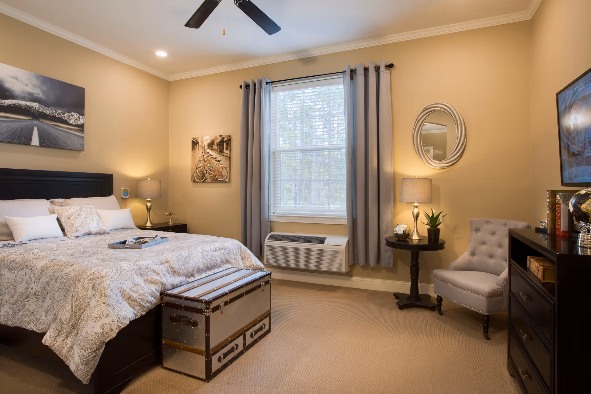 A cozy bedroom with a large bed featuring patterned bedding and multiple pillows. There is a decorative trunk at the foot of the bed. The room has beige walls, a window with gray curtains, a ceiling fan, two bedside lamps on nightstands, a round mirror on the wall, a small round table with a plant, a gray upholstered chair, and a dresser with books and a globe.