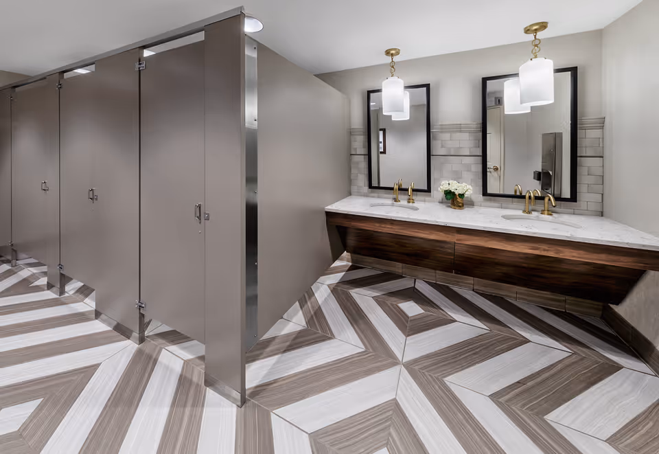 Modern public restroom with multiple toilet stalls, a double-sink vanity with mirrors and pendant lights, and a geometric patterned floor.