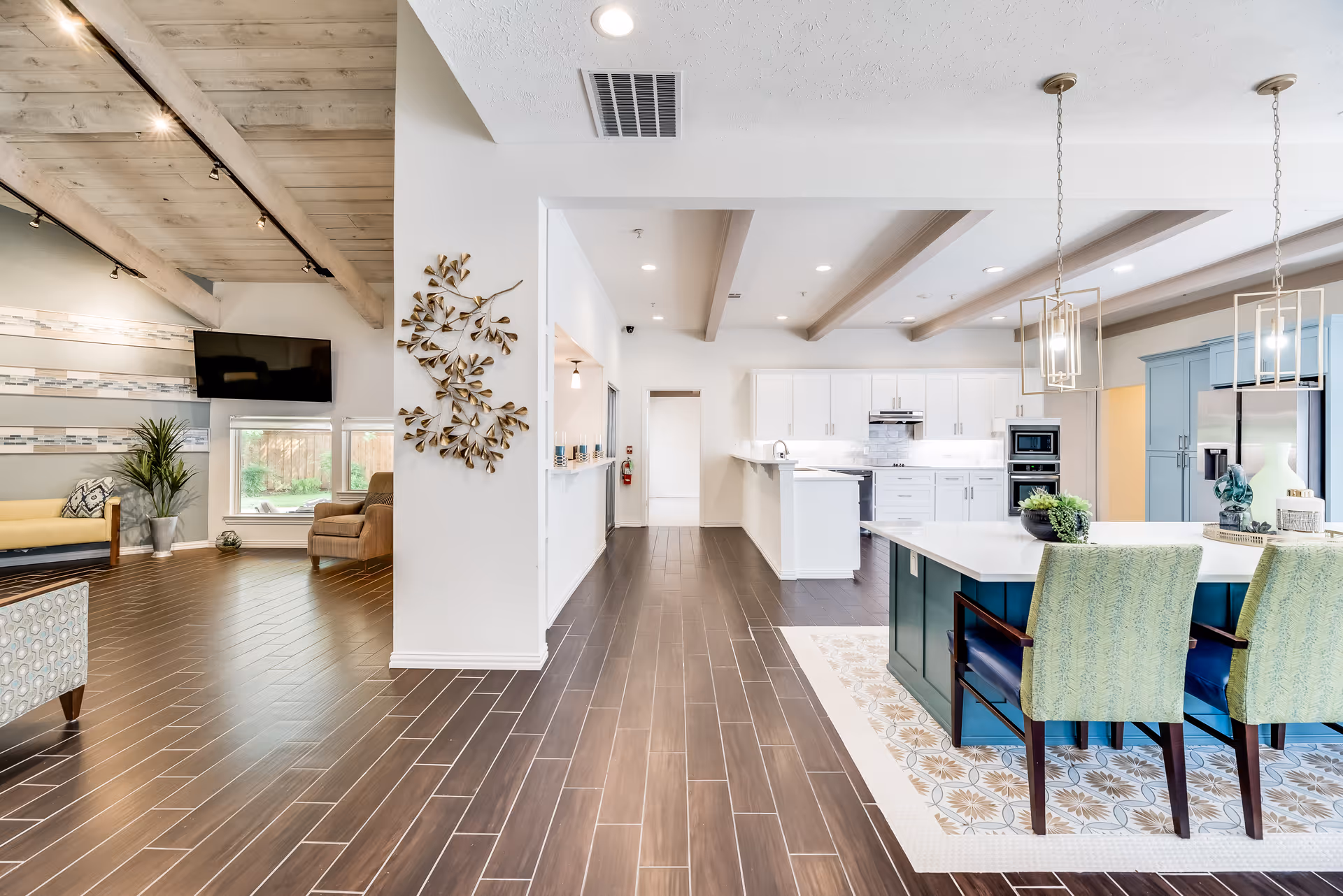 Open, bright interior common area showing a modern kitchen with an island and seating adjacent to a living room with chairs and a wall-mounted TV.
