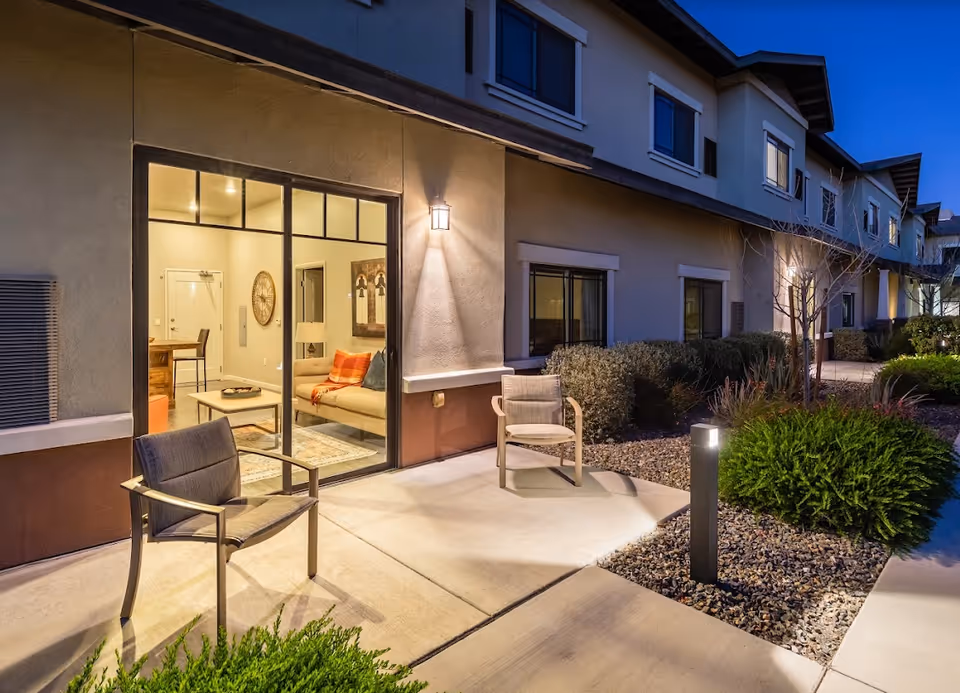 Evening view of an outdoor patio area with two chairs on a concrete surface next to a building. The building has large glass sliding doors revealing a cozy living room inside with a sofa, coffee table, and wall art. The patio is surrounded by small bushes and landscaping rocks, with pathway lighting and exterior wall lights illuminating the area.