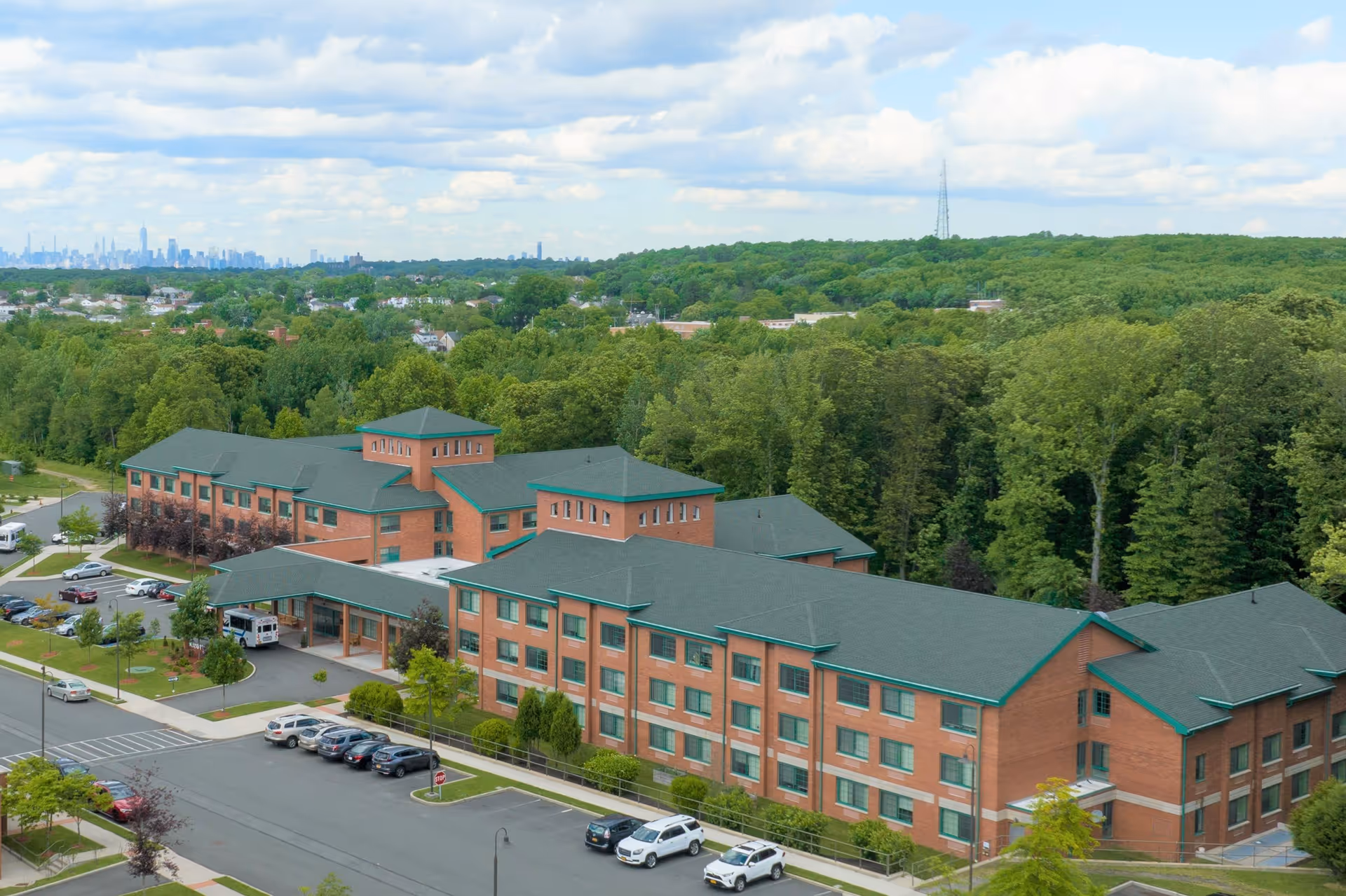 Aerial view of The Brielle senior living facility, a large multi-story brick building with green roofs surrounded by trees and parking lots with several cars parked. The background shows a distant city skyline under a partly cloudy sky.